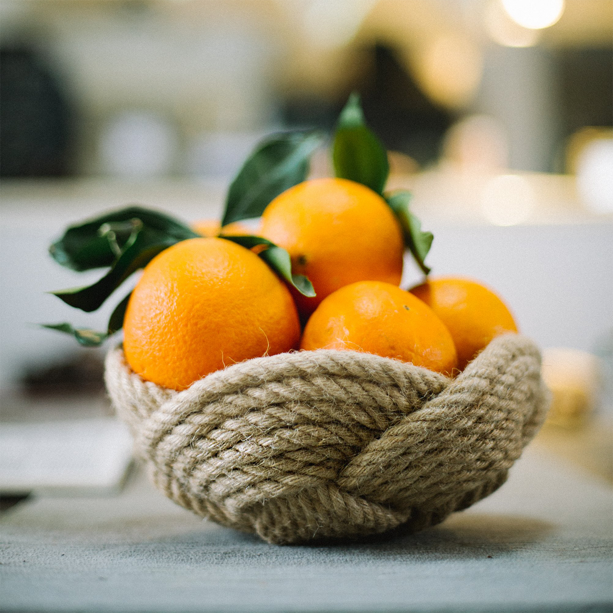Fresh oranges with green leaves in a woven rope basket on a table, natural light background