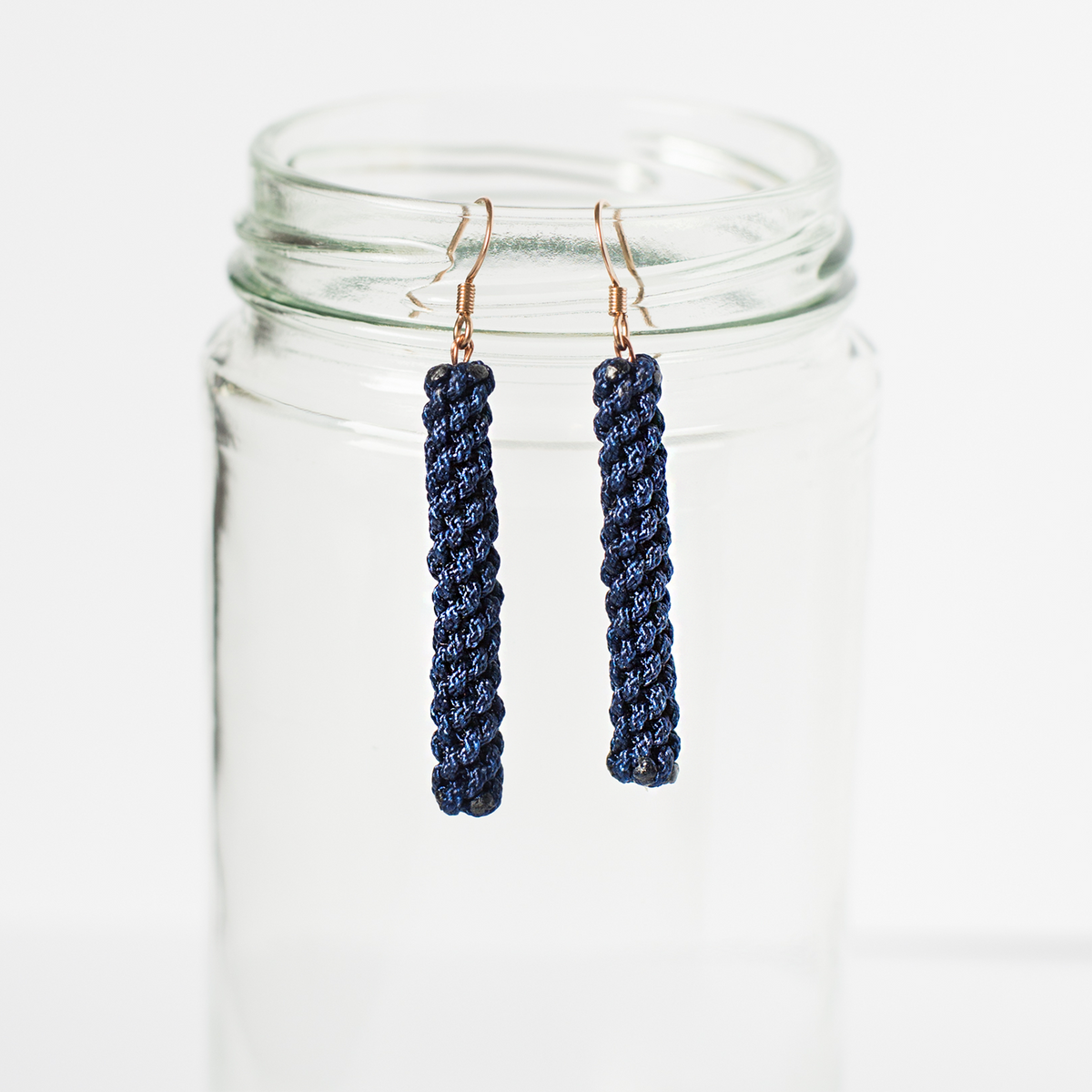 Handmade navy blue woven earrings with gold hooks displayed on a clear glass jar