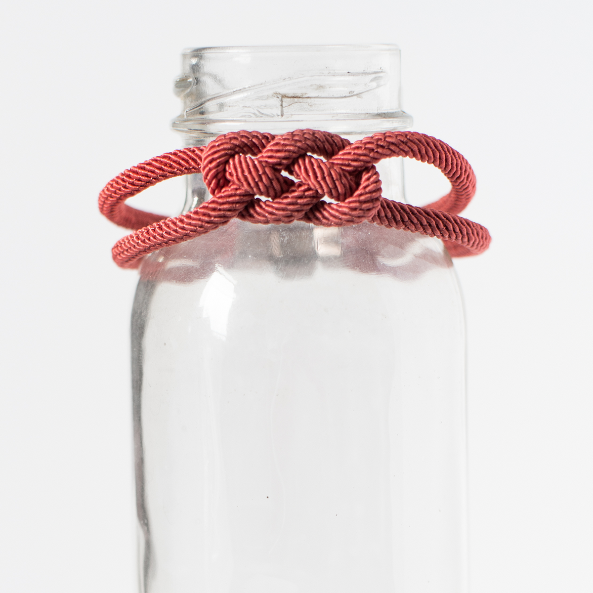 Red decorative rope knot bracelet displayed on clear glass bottle, minimalist background